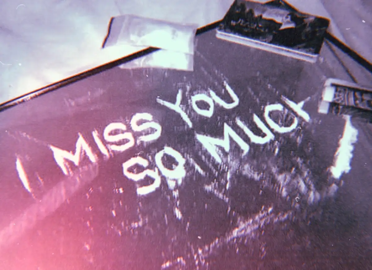 A close-up, grainy, vintage-style photo shows the words 'I miss you so much' written in white chalk or powder on a dark…