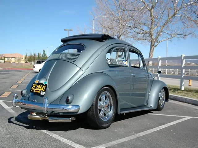 A custom-modified light blue Volkswagen Beetle is parked in an asphalt lot under a clear blue sky. The car features chrome…
