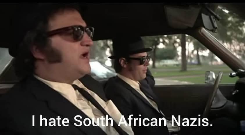 Two men in black suits, hats, and sunglasses sit in a car; the man in the passenger seat has his mouth open as if speaking…