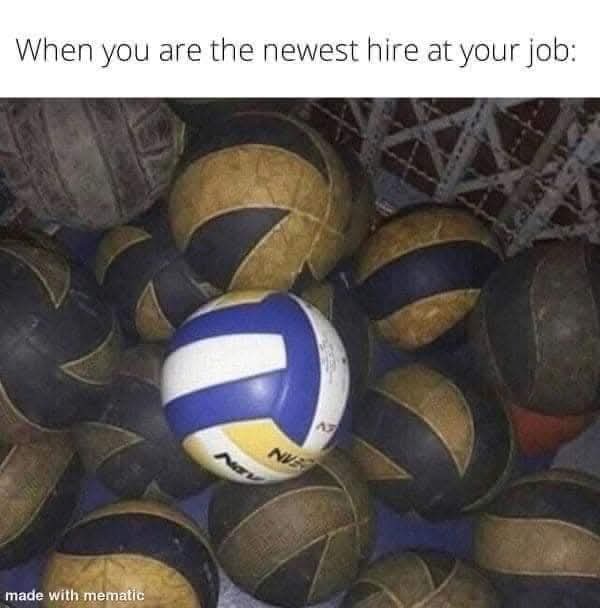 A close-up photo shows one bright, clean blue, white, and yellow volleyball sitting prominently among many worn, dirty, and…