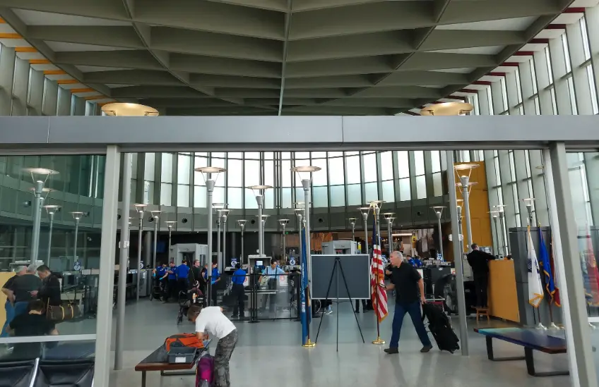 A wide shot of an airport security checkpoint area with travelers, security personnel, and flags. A man in a white shirt is…
