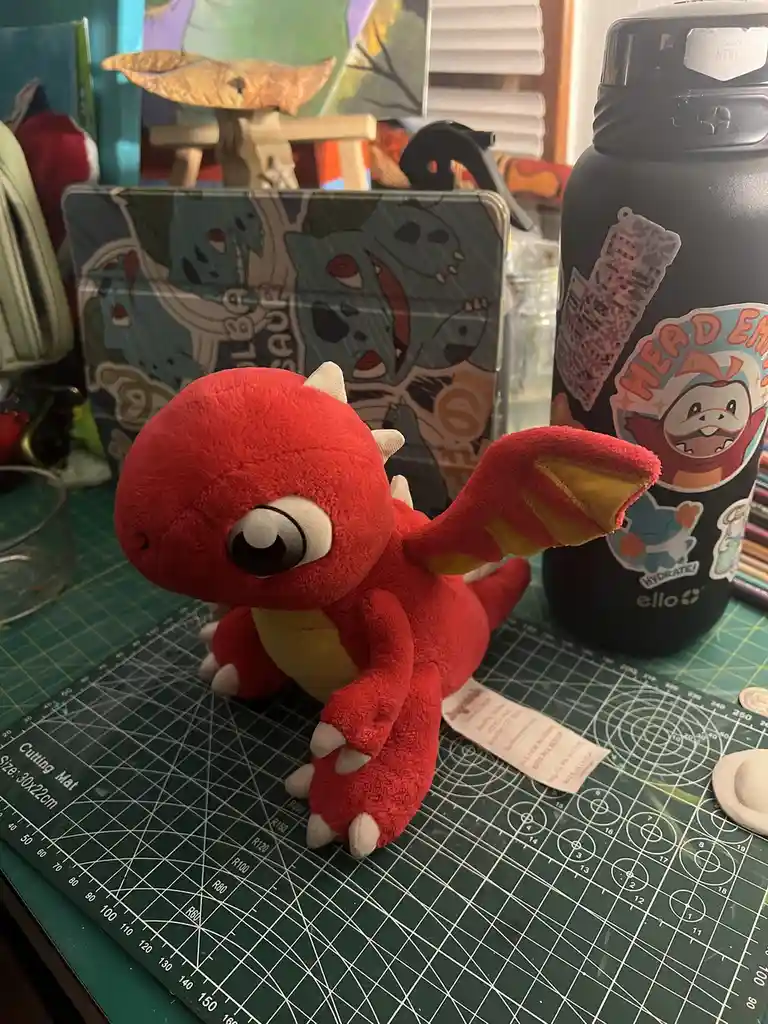 A small red dragon plushie with large eyes and white claws sits on a green cutting mat. Behind it is a tablet case with a…
