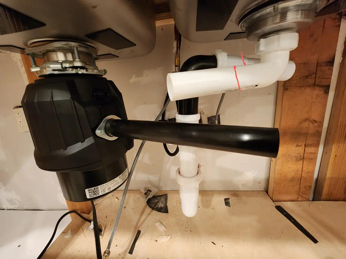 A photo under a kitchen sink shows a black garbage disposal unit connected to plumbing pipes, with a white PVC pipe…