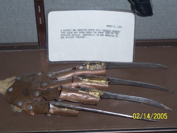 A rusted, broken Freddy Krueger-style glove with metal blades is displayed on a shelf next to a white plaque dated March 4…