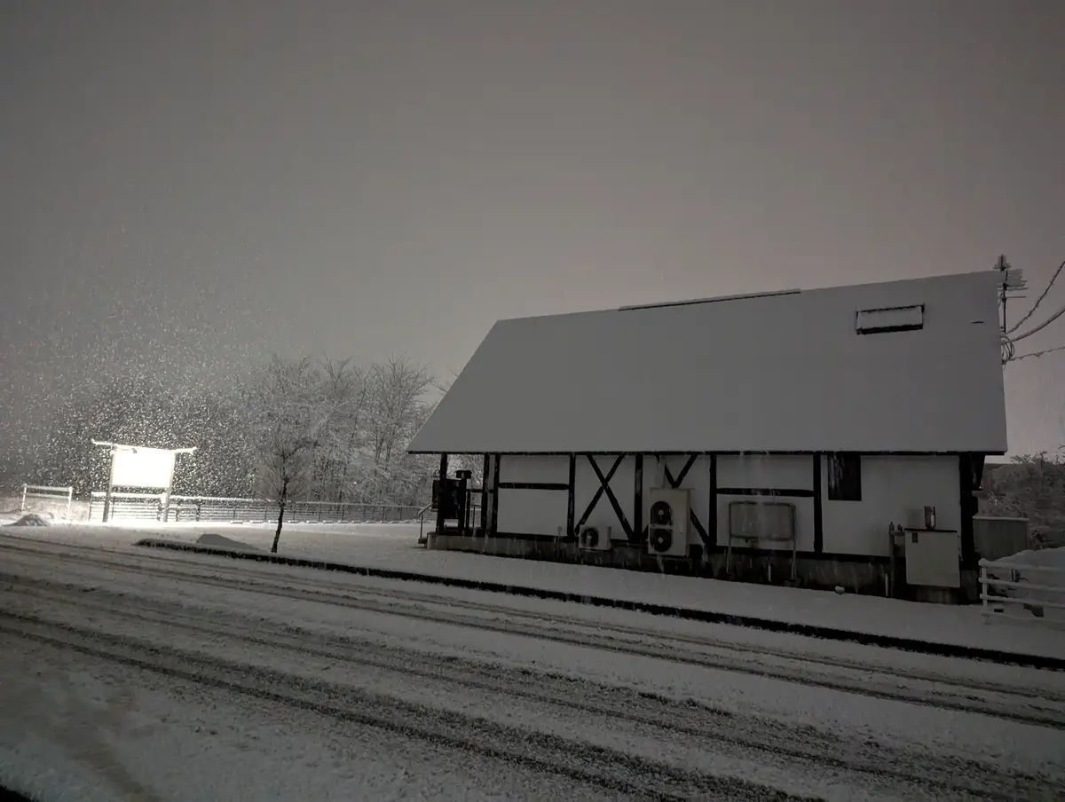 A snowy night scene shows a white building with black timber framing, its roof and ground covered in snow, with snowflakes…