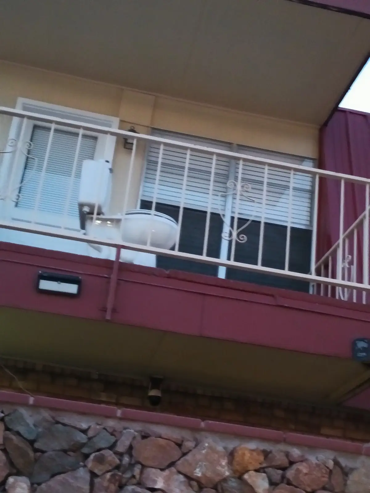 A white toilet is visible on a balcony behind a white metal railing, with windows and blinds in the background. The building…