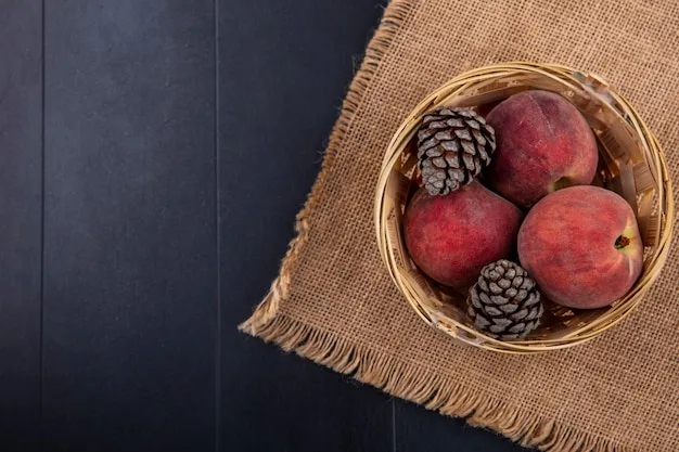 A top-down view of a woven basket containing several ripe peaches and two pinecones, resting on a burlap cloth over a dark…
