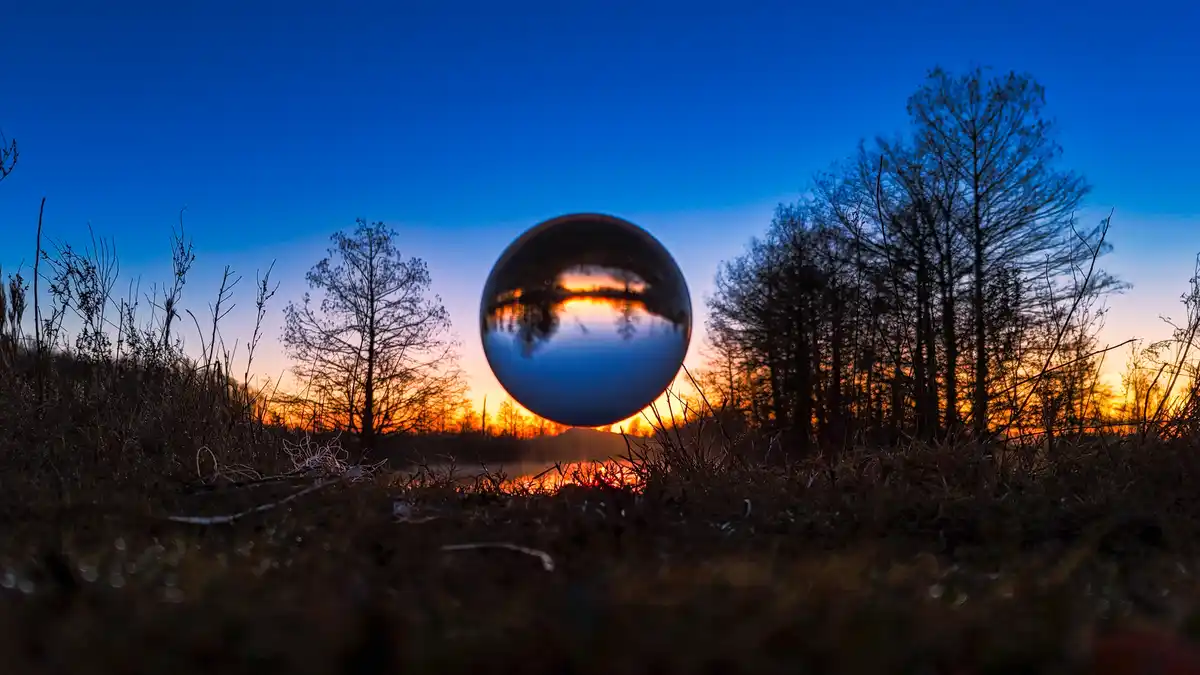 A reflective metal sphere sits on grass at sunset, capturing a distorted, inverted reflection of the sky and trees within…