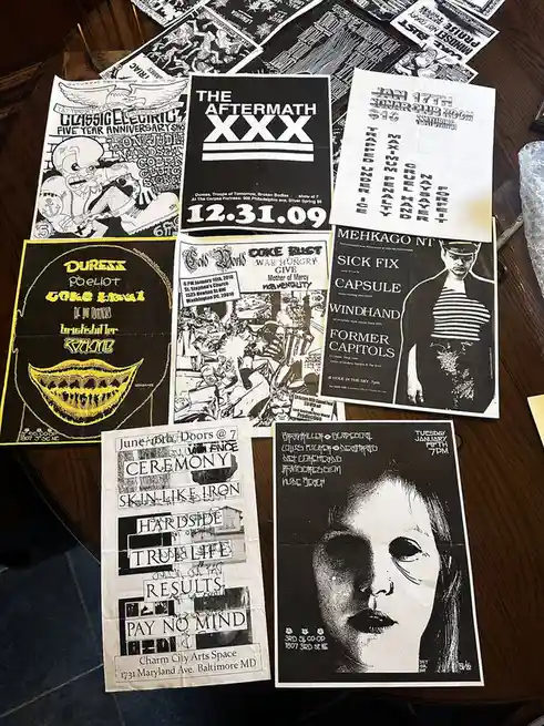 A collection of black-and-white punk and hardcore music flyers from 2009-2010 are scattered on a wooden table, featuring…
