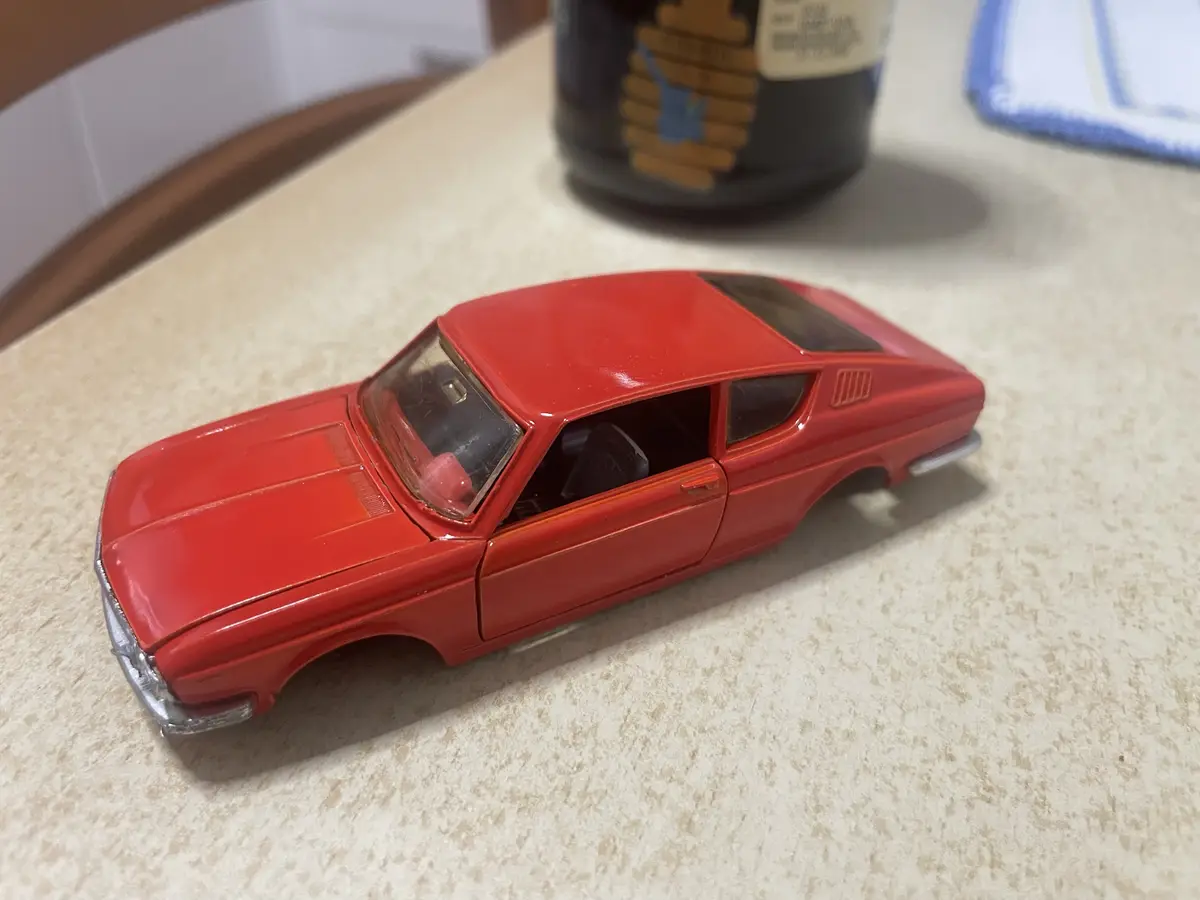 A red toy car with a glossy finish sits on a speckled countertop, its driver's side door open. In the background, a dark…