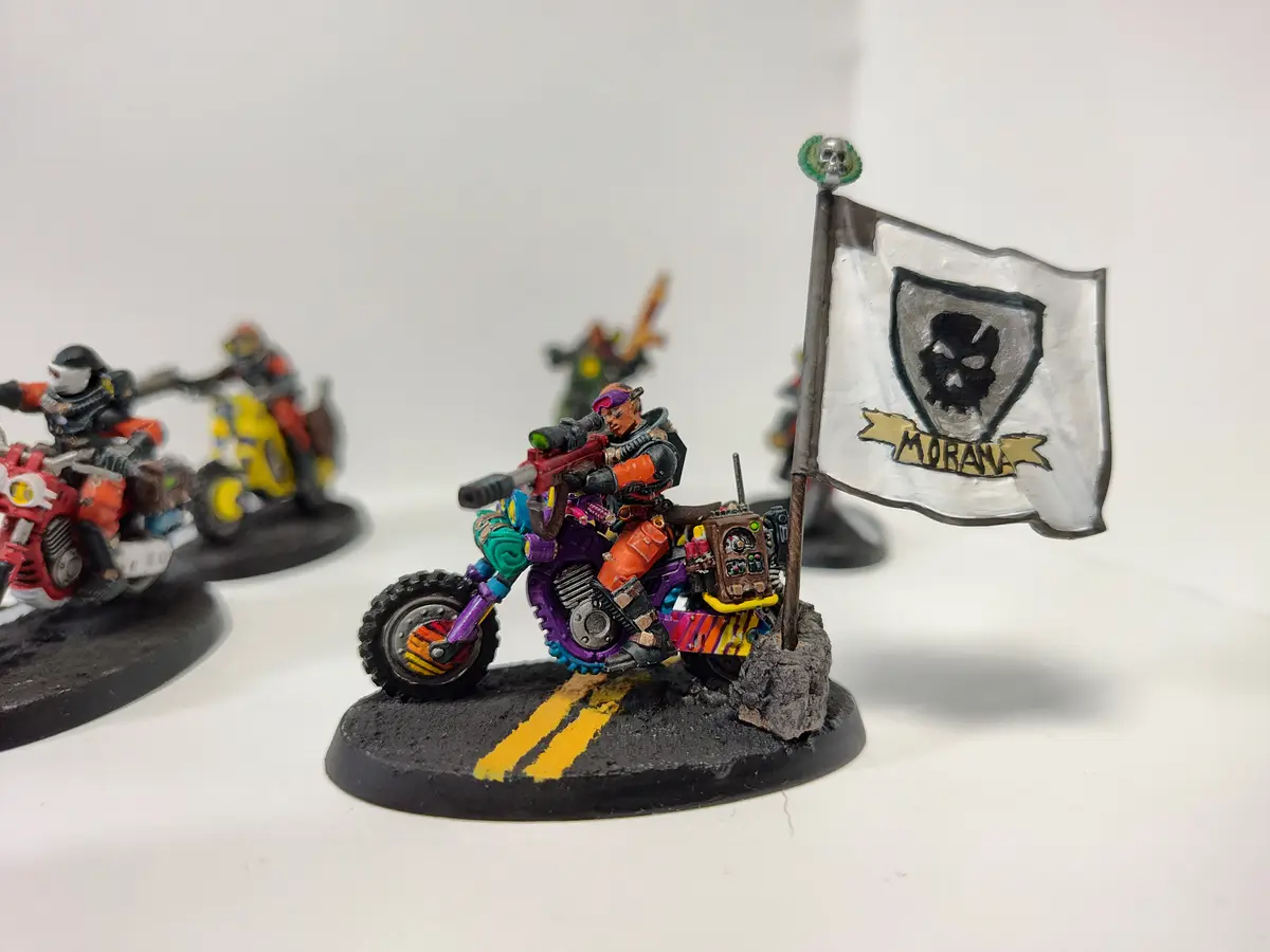 A painted miniature of a warrior on a colorful, heavily modified motorcycle, holding a flag with a skull emblem and the word…