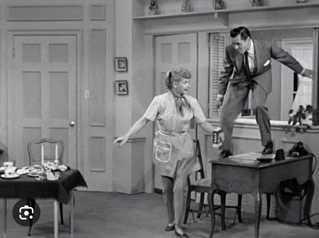 A black-and-white still from an old TV show shows a woman in an apron standing in a kitchen, looking alarmed as a man in a…