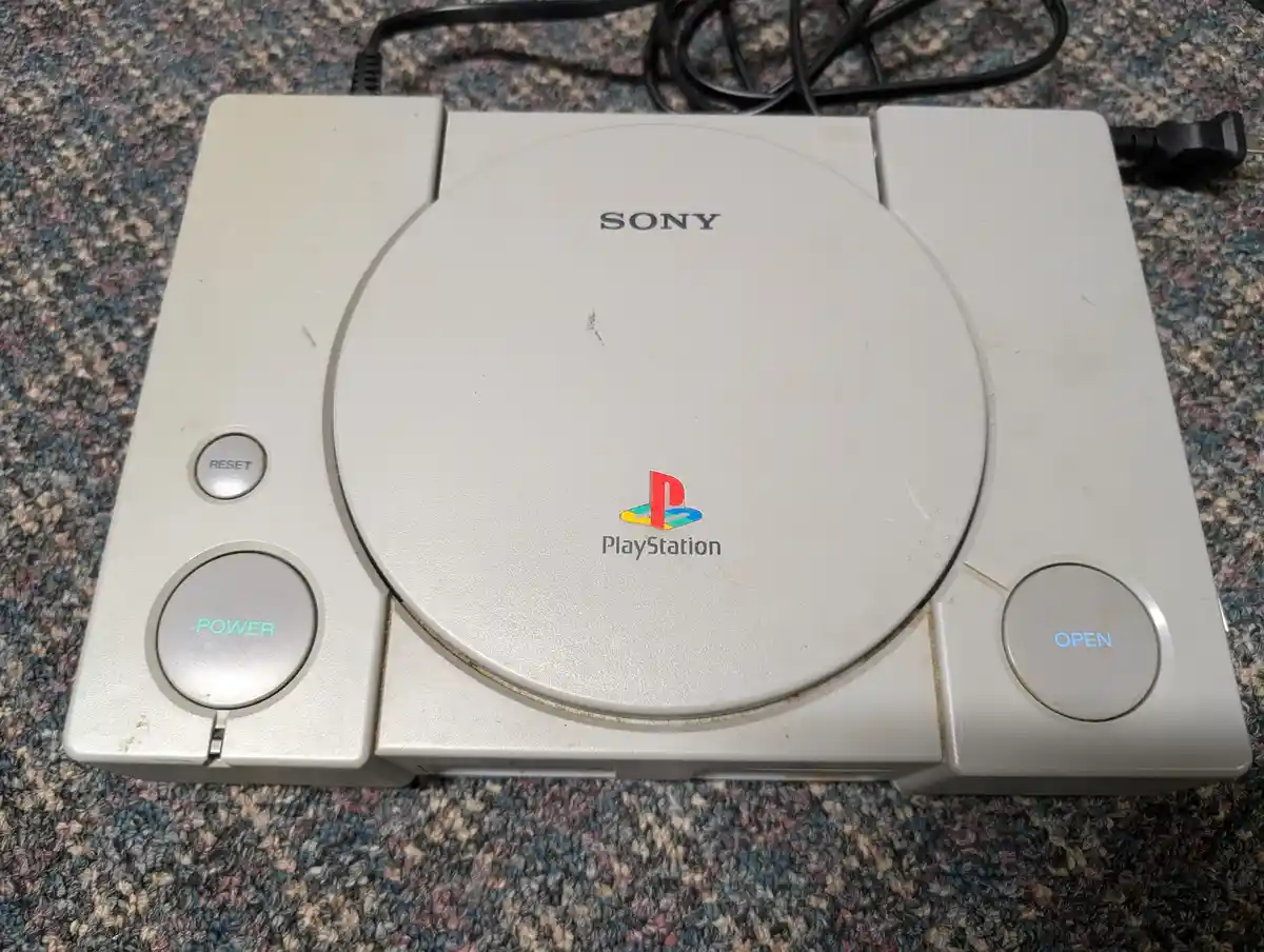 A top-down photo of a beige Sony PlayStation 1 console resting on a multicolored carpet, showing its circular disc slot…