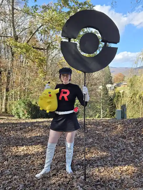 A person dressed as a Team Rocket member from Pokémon, wearing a black outfit with a red 'R' on the chest, white gloves, a…