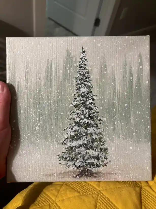 A hand holds up a small canvas painting depicting a snow-covered evergreen tree in a snowy forest, with visible brushstrokes…