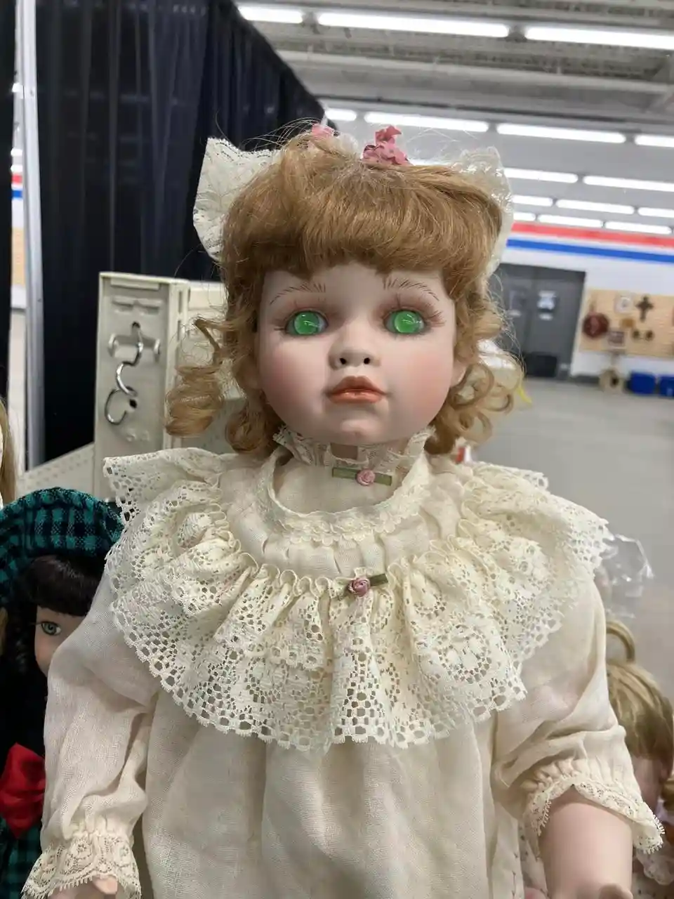 A close-up of a vintage-style doll with large, bright green eyes, curly brown hair, and white lace-trimmed clothing…