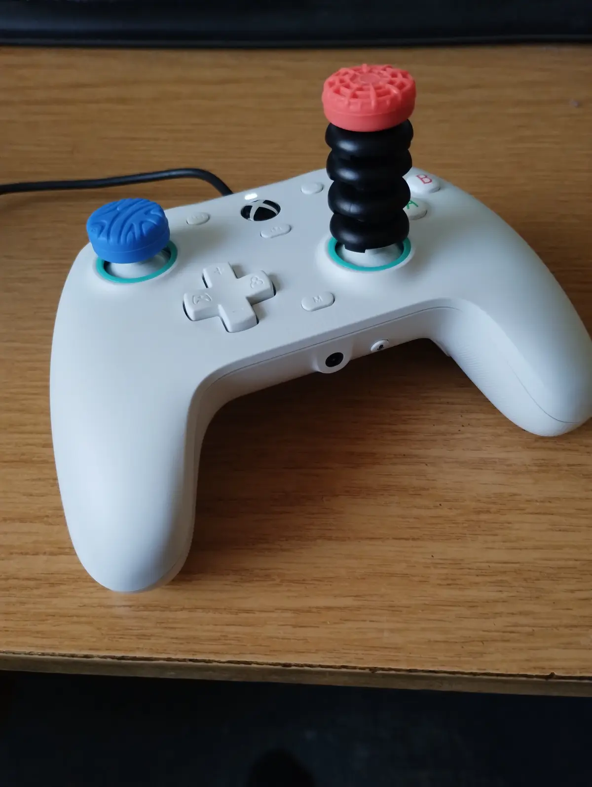 A white Xbox controller sits on a wooden surface, modified with a blue textured cap on the left stick and a tall, black…