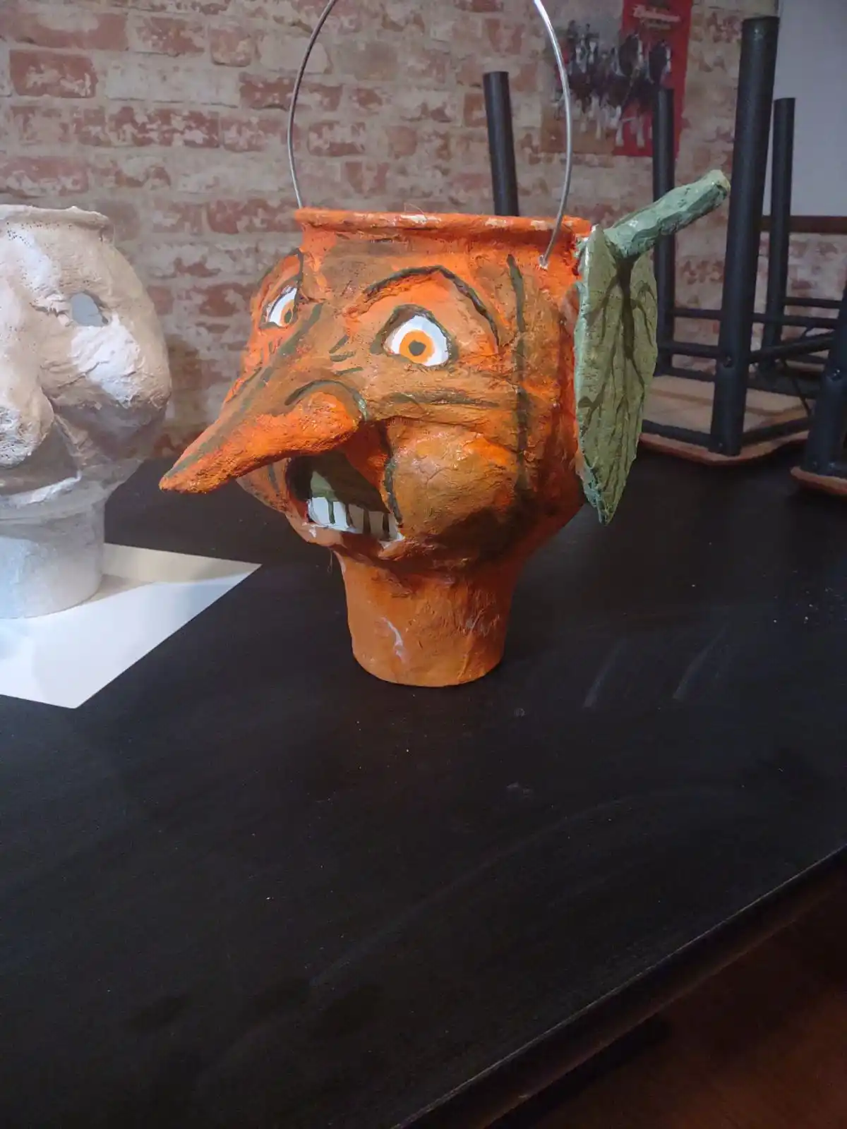 A handmade, orange, pumpkin-shaped bucket with a painted, screaming face, a long nose, and a green leafy ear, sits on a…