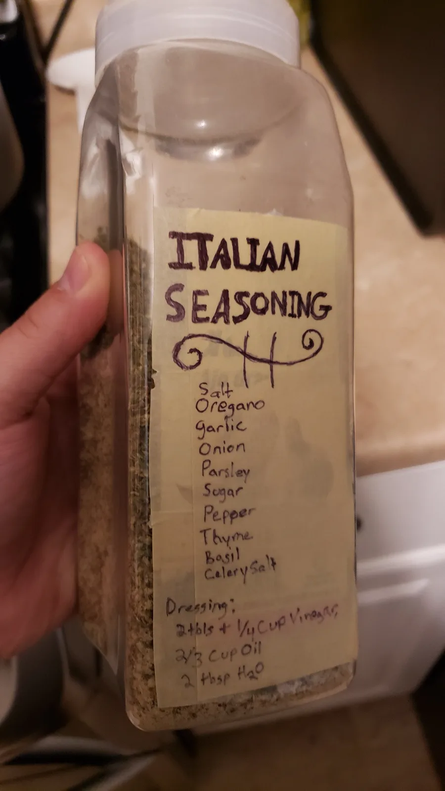 A hand holds a clear plastic spice bottle with a handwritten label titled 'ITALIAN SEASONING'. The label lists herbs and…