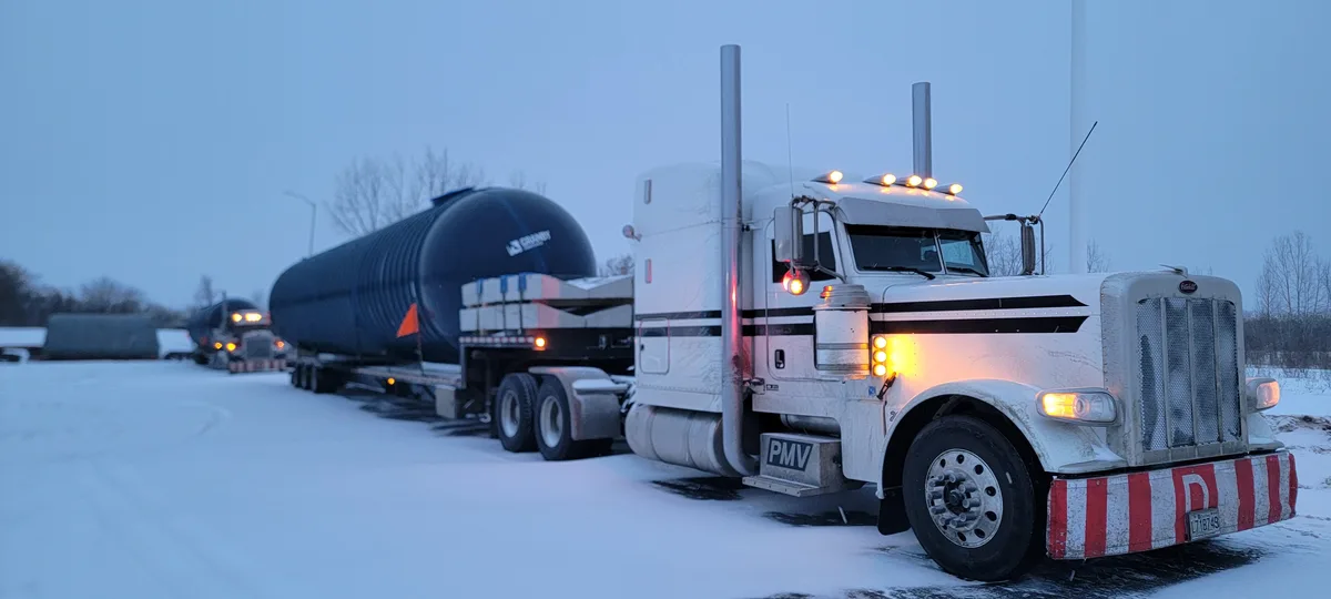 A white semi-truck with a large black tanker trailer is parked in a snowy lot during twilight. The truck's lights are on…