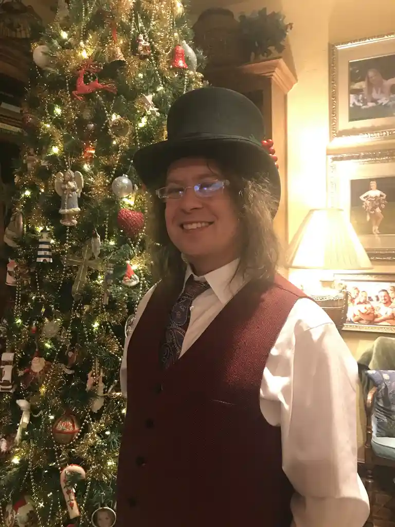 A person with long hair and glasses wears a black top hat decorated with red berries and a red vest over a white shirt and…