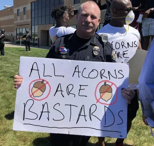 A police officer in uniform holds a handmade protest sign that reads 'ALL ACORNS ARE BASTARDS', with two hand-drawn acorns…