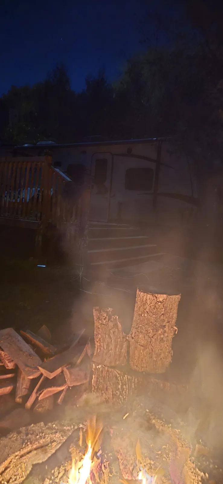 A nighttime photo shows a campfire burning in the foreground with smoke rising, surrounded by logs and firewood. Behind it…