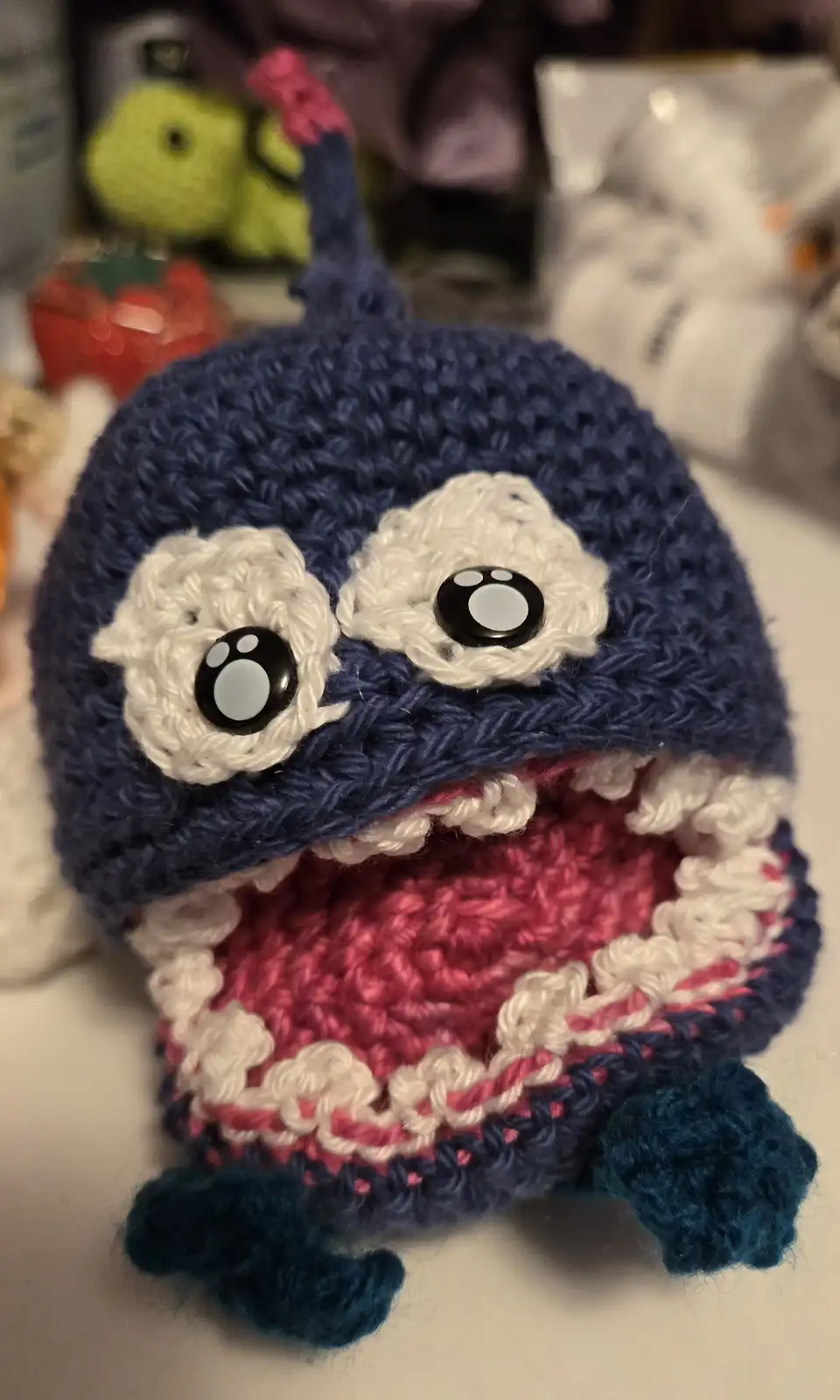 A close-up photo of a handmade crocheted plush toy resembling a cartoon shark with large white eyes, a wide open mouth…