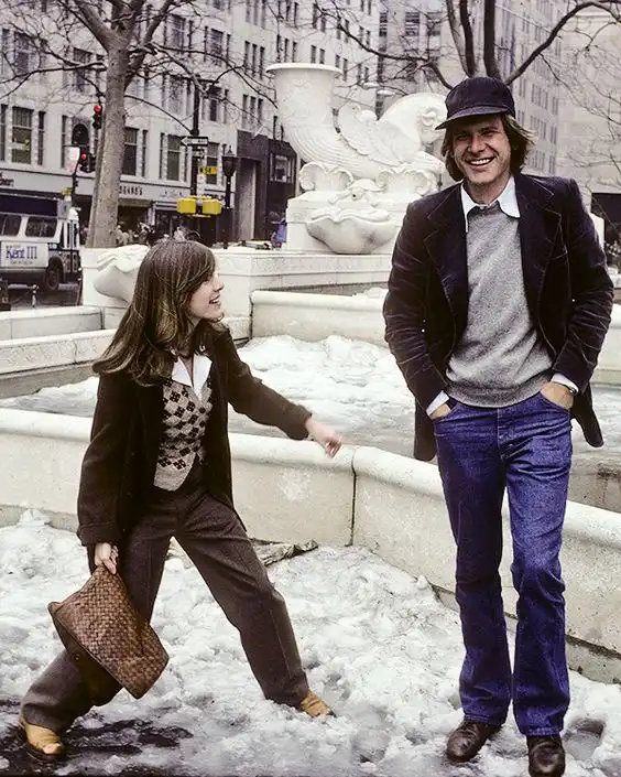A vintage photo shows a smiling man in a dark cap and velvet jacket standing beside a woman in a checkered vest and brown…