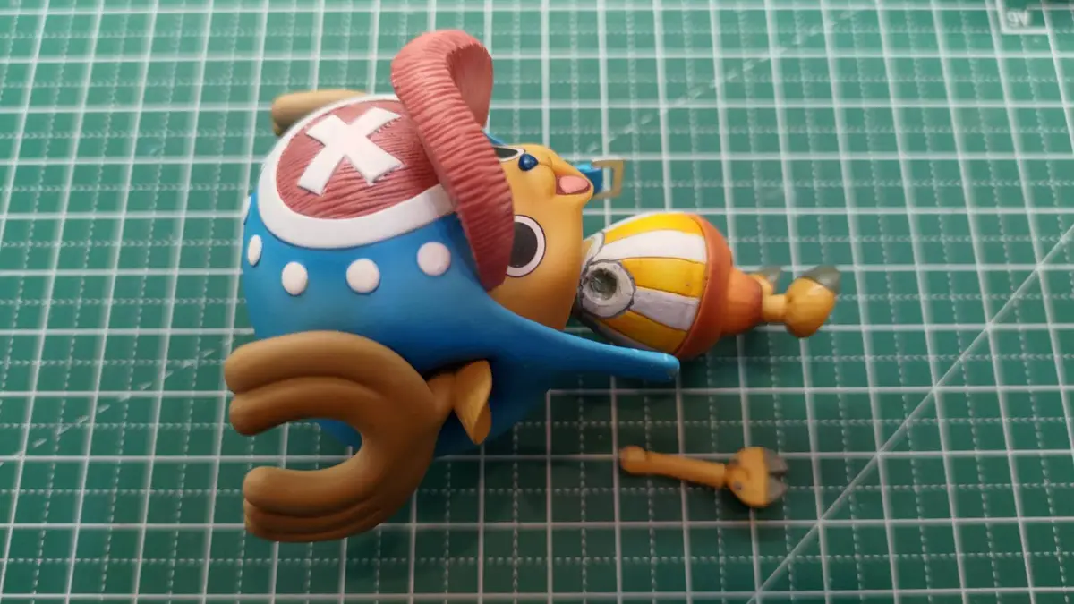 A broken One Piece figurine of Chopper is lying on a green cutting mat, with one arm detached and lying nearby. The figurine…