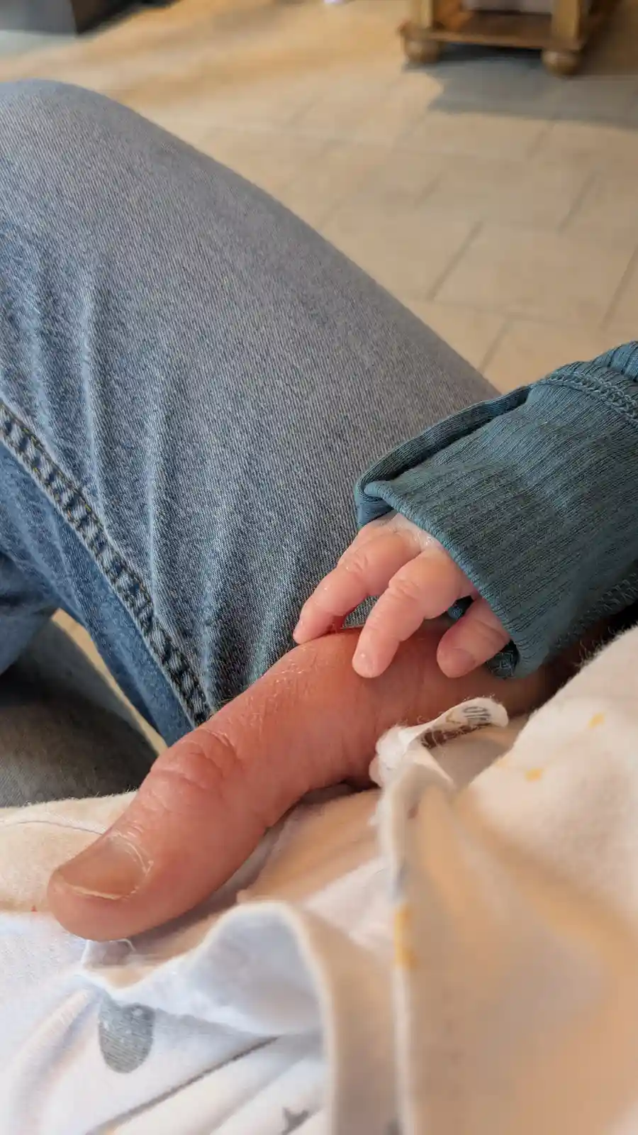 A close-up photo shows a tiny baby's hand gripping an adult's finger. The adult wears blue jeans and a teal long-sleeved…