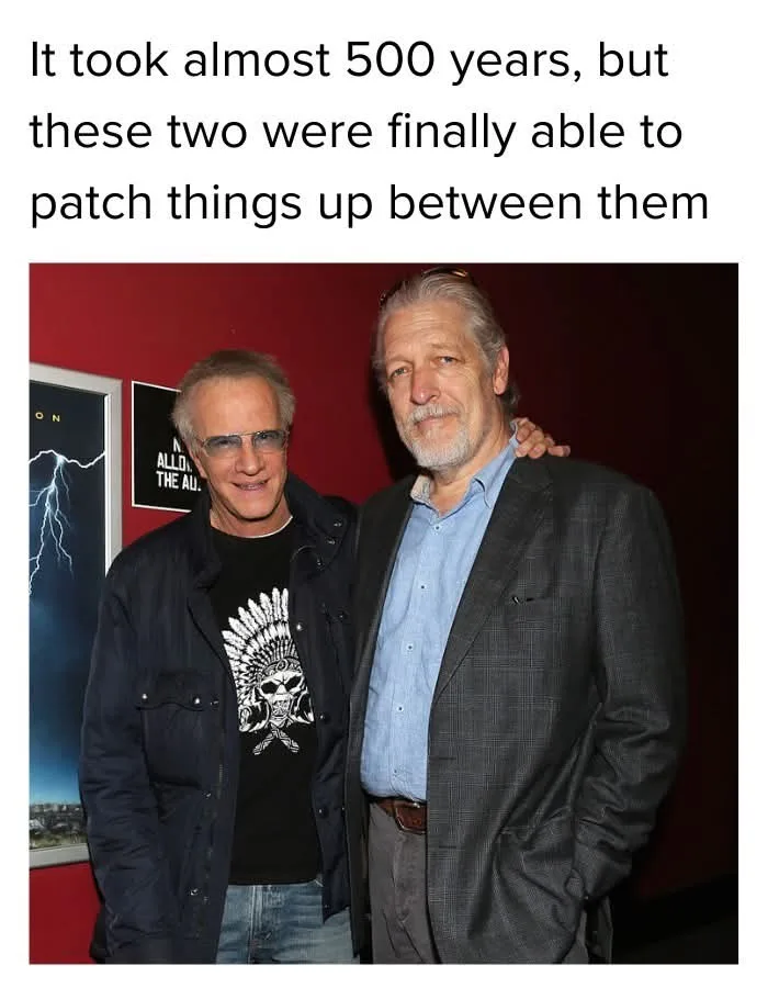 Two men, one in a black jacket over a skull-and-feather t-shirt and the other in a suit, pose together with friendly smiles…