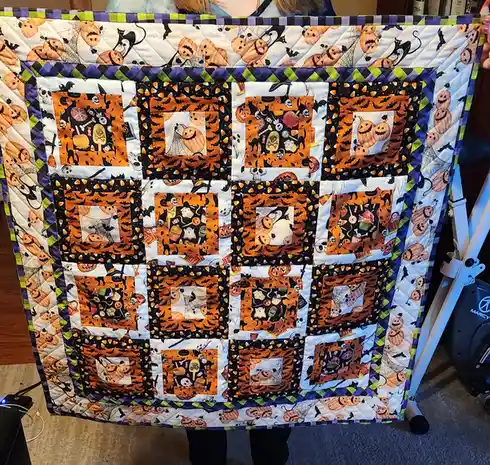 A person holds up a handmade Halloween-themed quilt featuring a grid of square patches with jack-o'-lanterns, bats, skulls…