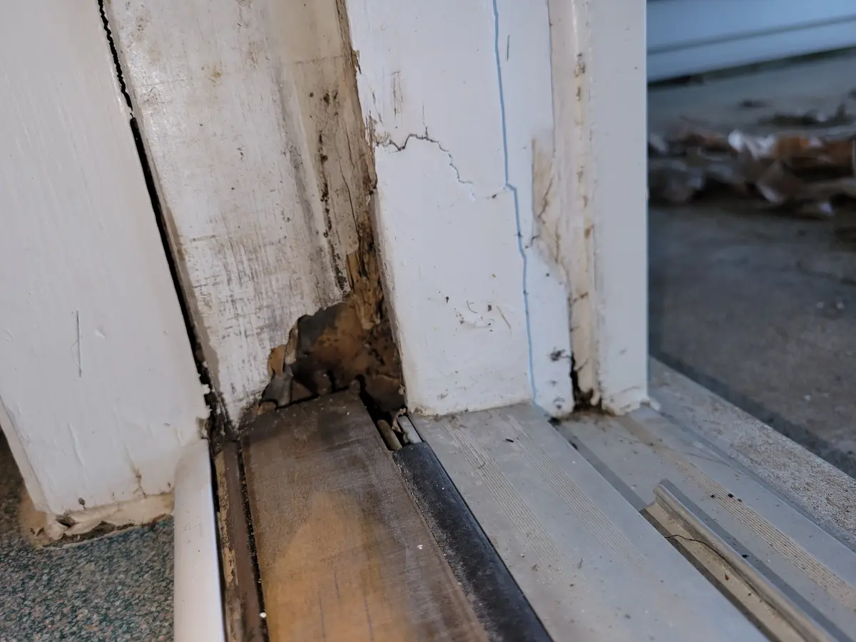 A close-up photo shows a severely damaged and rotting wooden door frame with peeling white paint, where the wood is…
