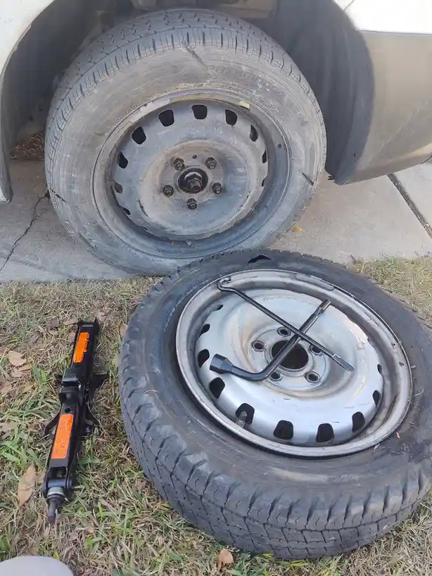 A flat tire with a silver rim and black lug wrench lies on grass next to a black car jack, while a spare tire with a gray…
