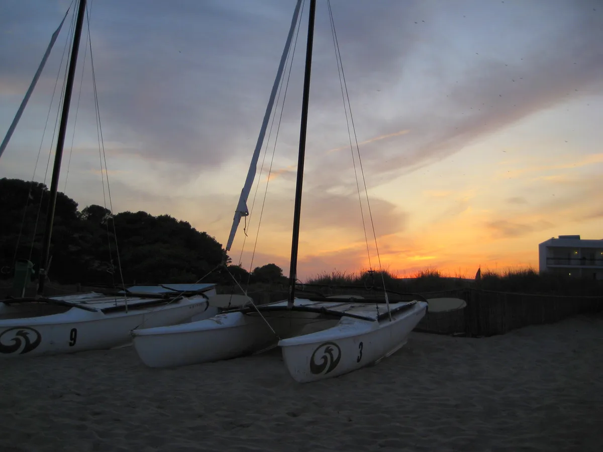 Two white catamaran sailboats sit on a sandy beach at sunset, their hulls marked with the number 3 and 9 and a swirl logo…