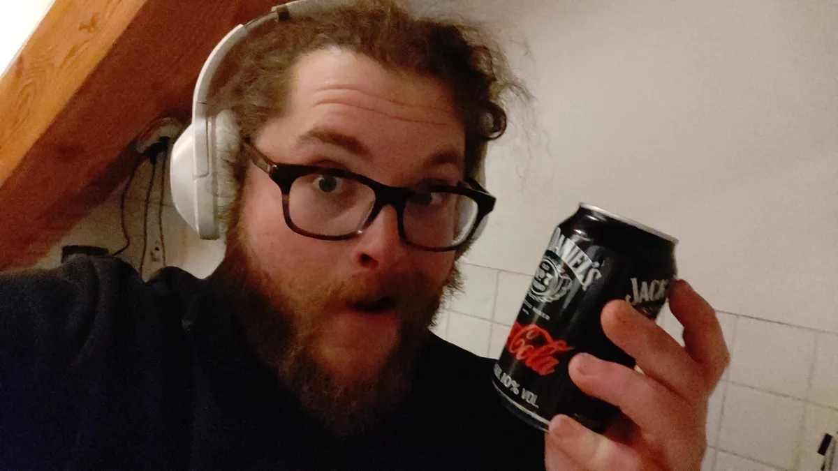 A man with a beard, glasses, and white headphones holds up a black can that has 'Jack Daniels' branding and 'Coca-Cola'…