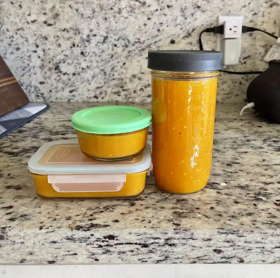 Three containers filled with orange-colored pureed soup sit on a speckled granite kitchen counter: a tall glass jar with a…