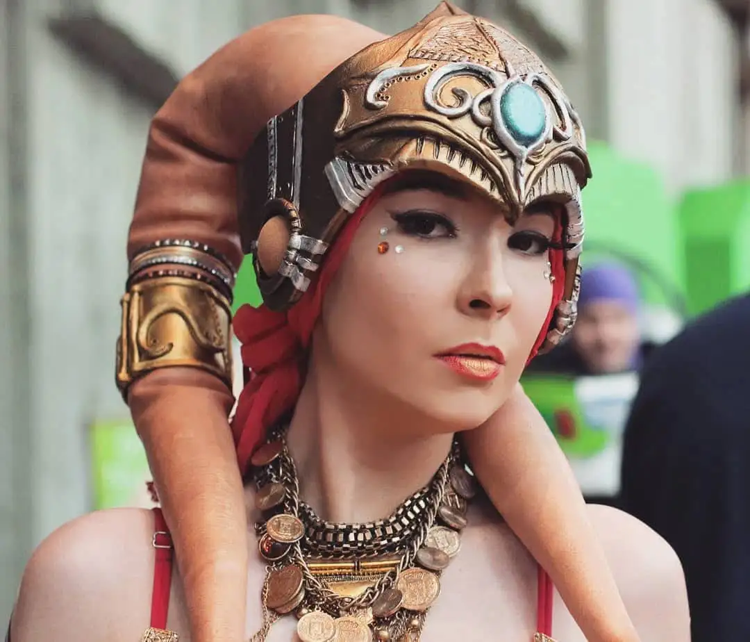 A person in elaborate fantasy cosplay, wearing a detailed golden helmet with a turquoise gem, red headscarf, and layered…