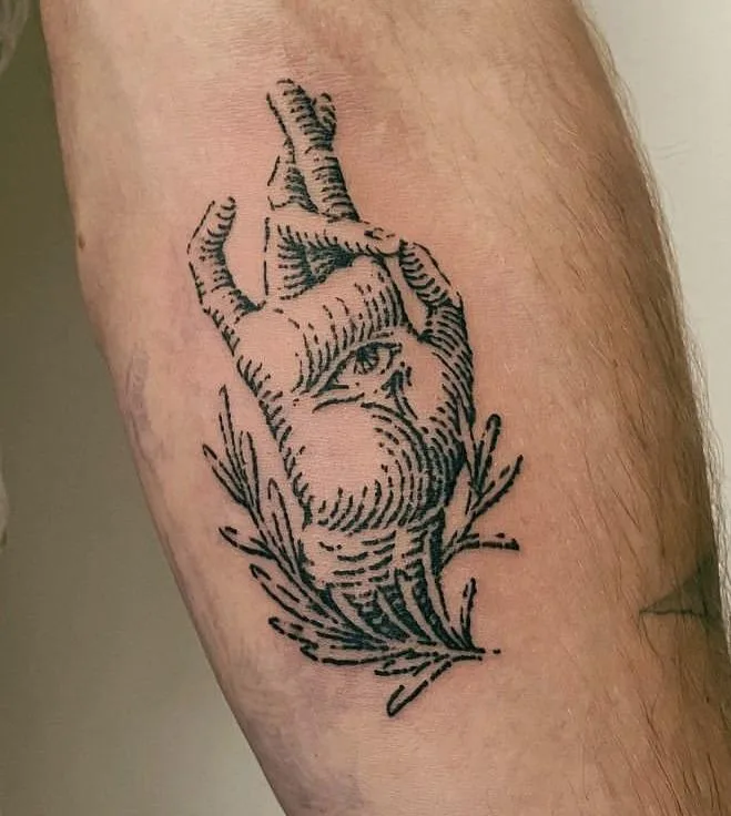 A black-and-white tattoo on a person's forearm depicts a hand with an eye in the center of the palm, framed by laurel…