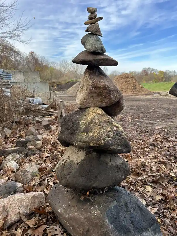 A tall, precarious stack of various-sized rocks is balanced outdoors on a bed of fallen brown leaves under a partly cloudy…