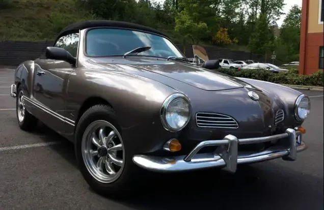 A dark gray vintage Volkswagen Karmann Ghia convertible is parked in a lot, featuring chrome bumpers and round headlights…