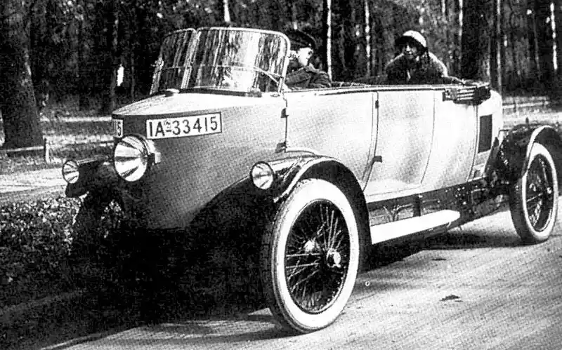 A black-and-white photograph of a vintage open-top automobile with two men inside, one driving and one in the passenger…