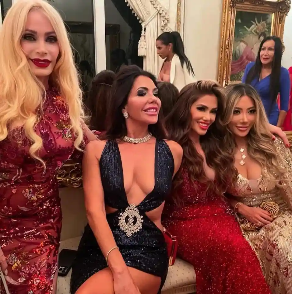 Four women in glamorous, sequined evening gowns pose together in a luxurious room with ornate decor and framed artwork. The…