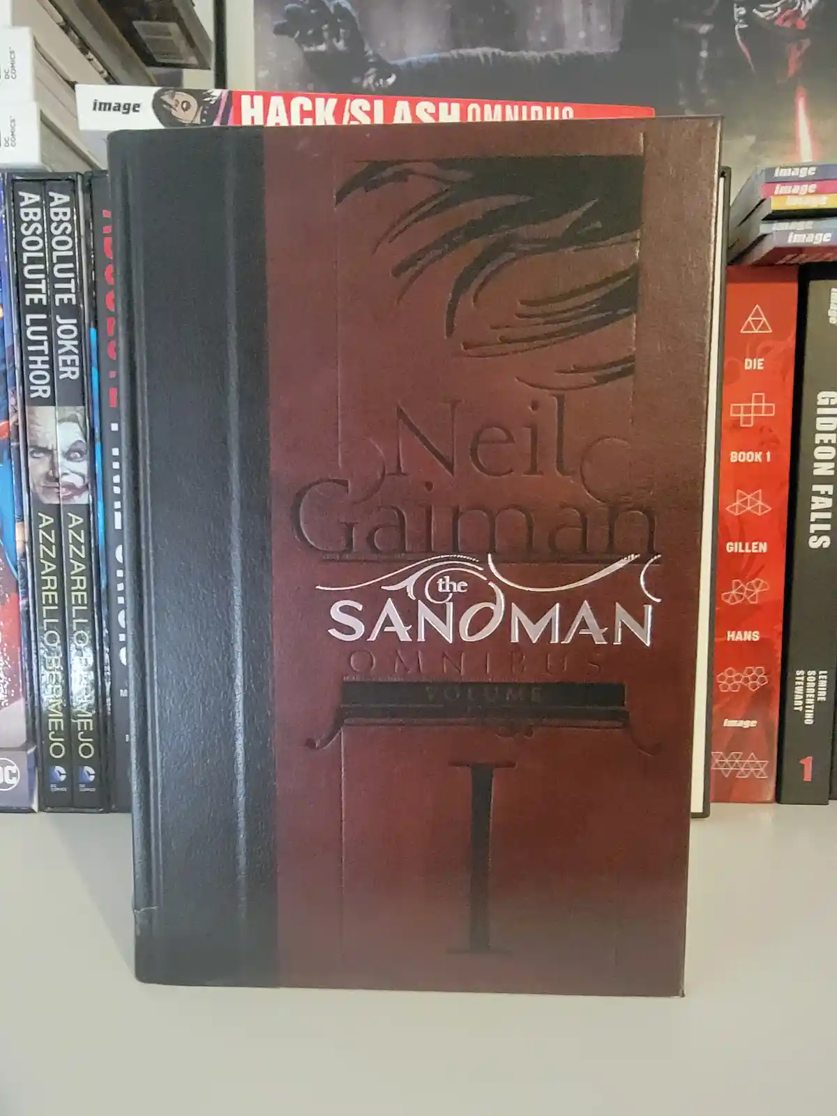 A large, dark brown leather-bound comic book titled 'Neil Gaiman the Sandman Omnibus Volume I' stands upright on a shelf…