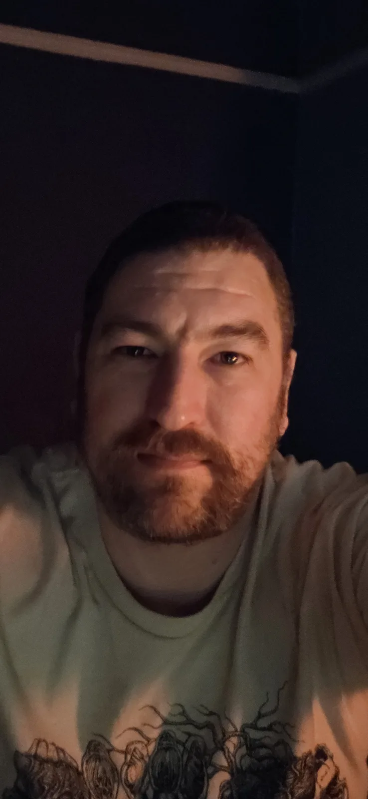 A close-up selfie of a man with a beard and short hair, wearing a light-colored t-shirt with a dark graphic, taken in dim…