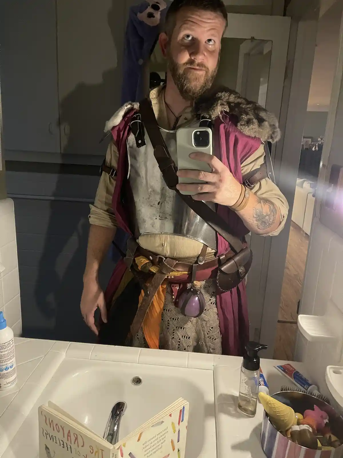 A man in a full fantasy warrior costume, complete with armor, fur-trimmed vest, and leather belts, takes a mirror selfie in…