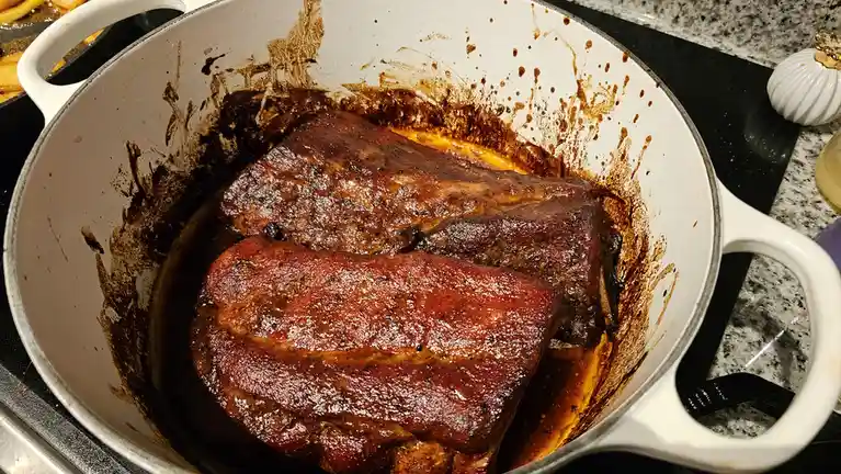 A white enameled cast iron Dutch oven sits on a stovetop, containing two large, glistening, glazed ribs that appear…