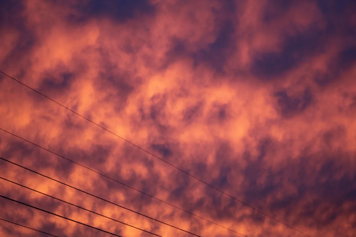 A dramatic sky filled with fiery orange and deep purple clouds, with several thin black power lines diagonally crossing the…
