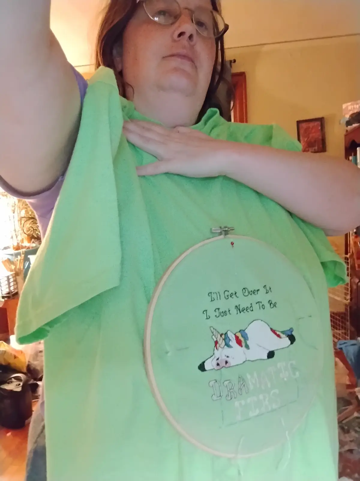 A person wearing a light green t-shirt holds up a large embroidery hoop displaying a stitched design. The design features a…